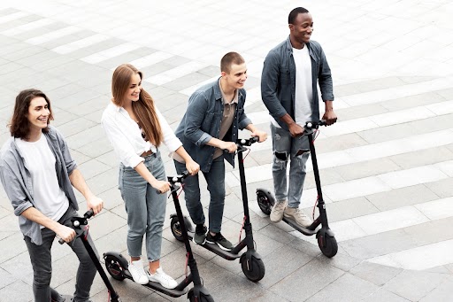 Four young adults smiling and walking together on a pedestrian plaza, each riding or standing with a black electric scooter. The group is casually dressed and appears to be in an urban outdoor setting with a crosswalk visible in the background.